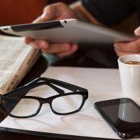 Newspapers and coffee cup, reading glasses, striped paper, hands holding tablet, cell phone.